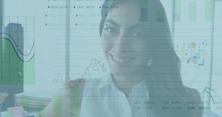 Sitting woman wearing white collared blouse studying overlaid charts at office desk, with monitor