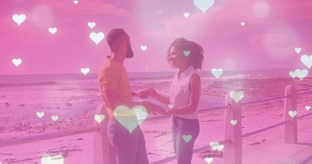 Holding hands couple in casual outfits smiling and leaning on promenade, railing and heart bokeh