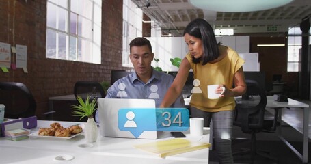 Pointing coworkers in mustard blouse and blue shirt leaning over laptop, reviewing alert at office