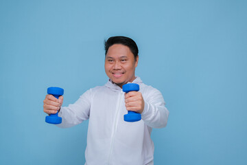 Close up of young Asian man in the gym lifting dumbbell wearing white t-shirt with happy laughing expression on turquoise blue background