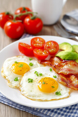 Delicious breakfast plate includes sunny-side-up eggs, crispy bacon, vibrant tomatoes, and creamy avocado, presented on a white dish with a soft gray napkin