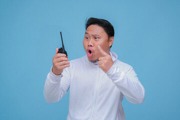 Young Asian man talking on Handy Talkie with very angry expression wearing white t-shirt on turquoise blue backgroun