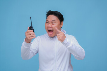 Young Asian man talking on Handy Talkie with very angry expression wearing white t-shirt on turquoise blue backgroun