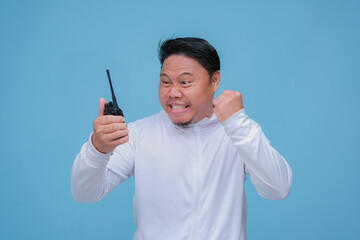 Young Asian man talking on Handy Talkie with very angry expression wearing white t-shirt on turquoise blue backgroun