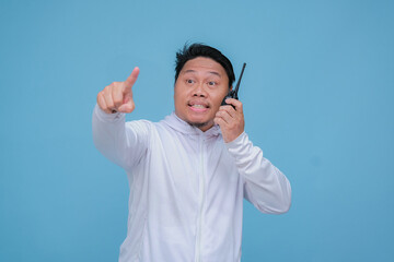 Young Asian man talking on Handy Talkie with very angry expression wearing white t-shirt on turquoise blue backgroun