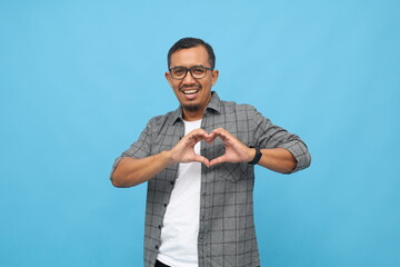 Photo of smiling asian man isolated on blue studio background, showing heart-shaped hand gesture, showing sign of love, sharing affection and care.