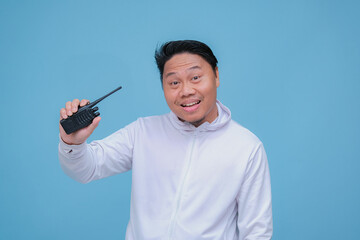 Closeup of Asian man talking on Handy Talky with laughing expression and pointing finger wearing white t-shirt on turquoise blue background 