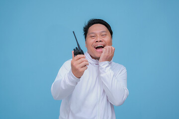 Closeup of Asian man talking on Handy Talky with laughing expression and pointing finger wearing white t-shirt on turquoise blue background 