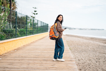 Smiling adult woman with dark hair wearing casual clothes and orange backpack holding smartphone while standing on wooden beach promenade near sandy shore and calm sea.