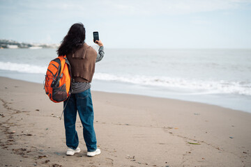 Woman with backpack taking smartphone photo on sandy beach near ocean waves, modern travel lifestyle, digital nomad concept, freedom, technology, seaside environment, remote communication
