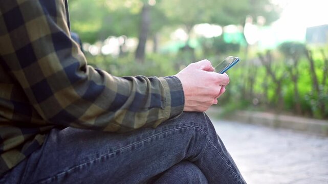 Vertical footage shows a man wearing gray jeans and plaid green shirt using his mobile phone outdoors. Selective focus, nice soft background.