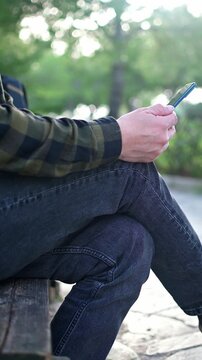Lockdown static shot footage  shows a man wearing gray jeans and plaid green shirt using his mobile phone outdoors. Selective focus, nice soft background.
