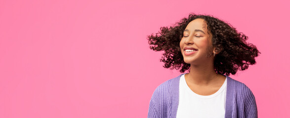 A young woman with curly hair is smiling and standing against a bright pink background. She looks...