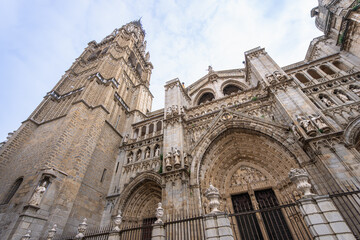 Gothic facade of the Primatial Cathedral of Saint Mary of Toledo featuring intricate stone carvings and a tall bell tower under a bright sky.