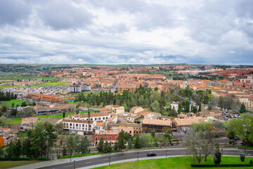 A scenic overview of Toledo, Spain, featuring historic architecture, green spring trees, and a cloudy sky above the ancient city landscape.
