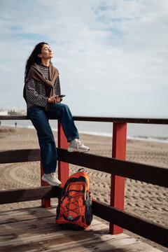 Relaxed adult woman sitting on wooden beach pier with backpack and smartphone, eyes closed, casual clothing, peaceful seaside atmosphere, outdoor lifestyle moment, freedom and mindfulness
