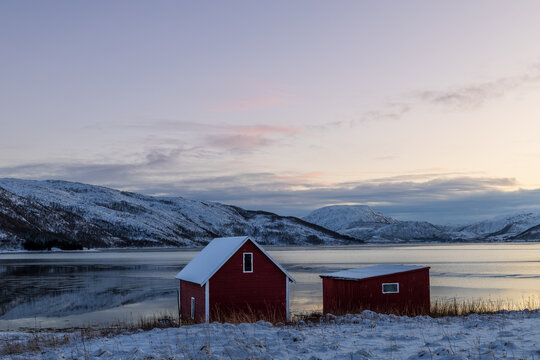 Zwei kleine norwegische Holzh&uuml;tten am ruhigen See in winterlicher Landschaft, schneebedeckte Ufer und Berge im Hintergrund, klare Abendstimmung mit sanftem Restlicht am Himmel 