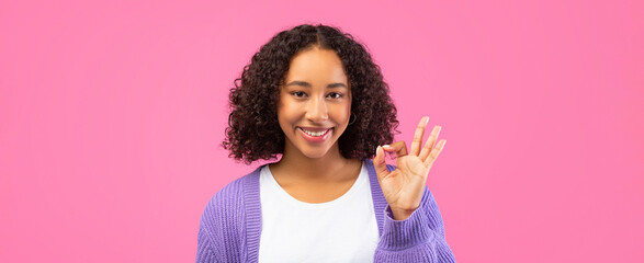 A young woman stands against a pink background. She smiles while making an okay sign with her right hand. Her curly hair frames her face. She wears a simple white top and a purple sweater.