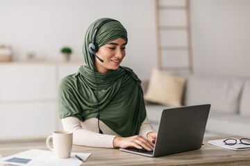 A millennial Arab lady in a green hijab is using her laptop and wearing headphones. She is working remotely from home and actively communicating with a client during an online meeting.