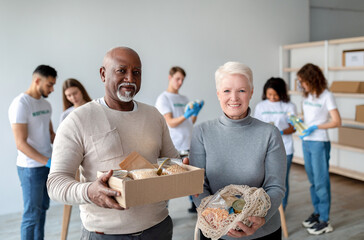 A happy mixed race senior couple smiles at the camera while holding a donation box and string bag...