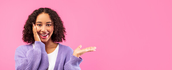 A young girl with curly hair smiles widely while resting one hand on her cheek. She gestures with her other hand, creating an inviting and cheerful moment in front of a pink wall.