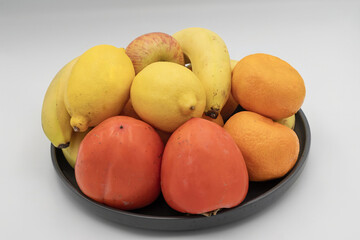 Healthy fruit mix in dark bowl with bananas citrus apples and persimmons on white studio background