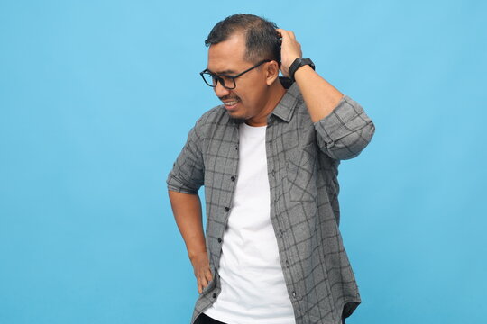 Asian man looking down holding his head, feeling disappointed, experiencing failure, ashamed of making a mistake, standing isolated on blue background.