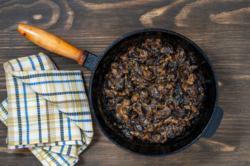 Cast iron skillet with fried wild mushrooms with onions and sour cream on wooden background, top view, closeup