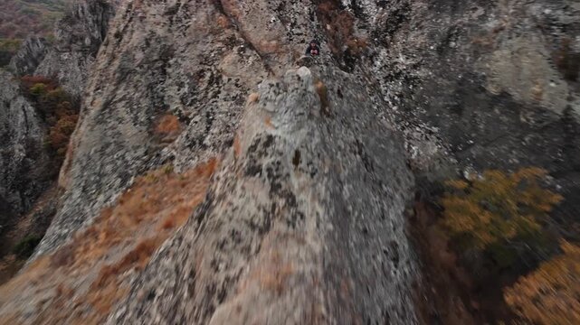 Man on cliff controls drone flying off into distance, showing mastabas of Georgia mountains and natural landscapes, overcast sky, physical exertion and focus, technical route on rugged terrain.