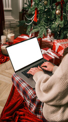 Person typing on laptop near Christmas tree and gifts, cozy holiday atmosphere