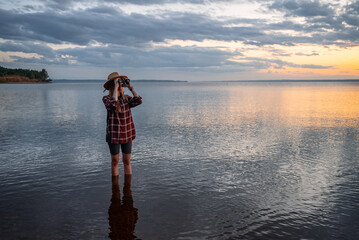 Woman standing in lake water at sunset, wearing a hat and plaid shirt, observing the vast natural landscape with binoculars, embodying exploration, freedom, and tranquil contemplation