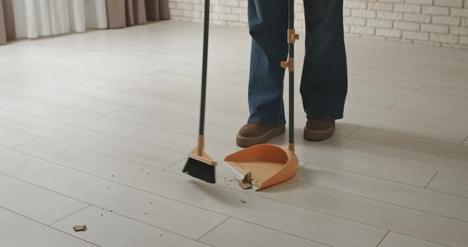 A person in jeans and shoes sweeping a light wood floor with a broom and dustpan, capturing a cleaning moment. Slow motion.