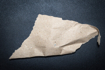 A torn piece of brown paper lies on a black textured surface, showcasing its rough edges and natural fibers. Ideal for backgrounds in design projects or craft materials.