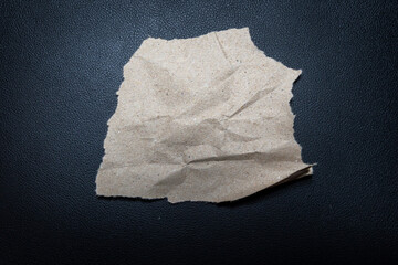 Crumpled piece of brown recycled paper placed on a textured black surface. Ideal for concepts of waste, recycling, environment, or craft projects.