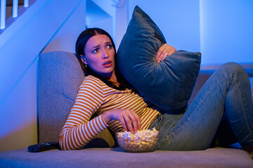 Scared woman watching terror movie in the night, sitting on couch with bowl of popcorn and hugging pillow in fear