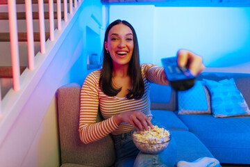 Excited woman sitting on sofa, eating popcorn, switching channels with remote controller, watching TV at home in the evening