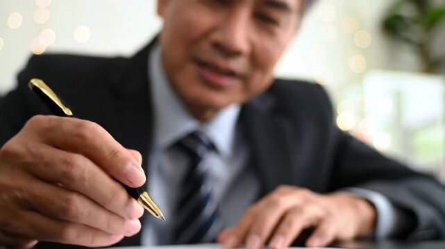 Senior Asian man in suit signing a document with pen, writing on paper. Focus on hand, business concept.
