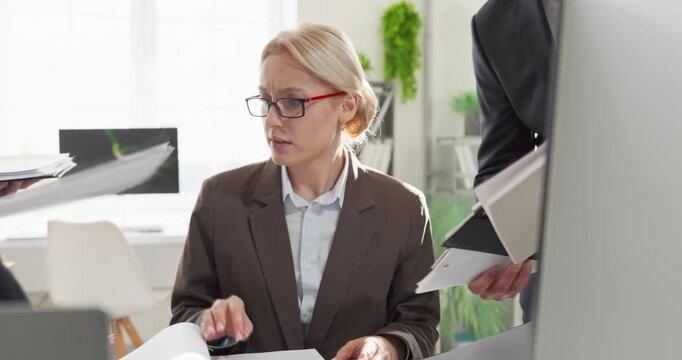 Businesswoman under stress in office facing deadline. Overwhelmed woman juggles documents from coworker at desk with heavy workload and time pressure. Clear concept of workplace burnout.