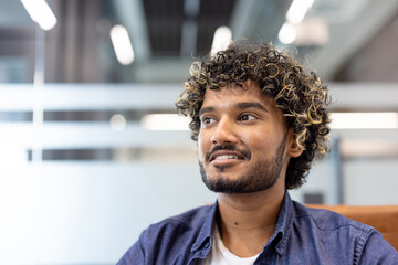 Close-up photo of a young Indian man sitting in an office and looking to the side with a smile
