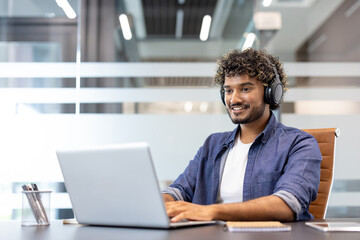 Smiling young Indian man in headphones working and studying in the office on a laptop
