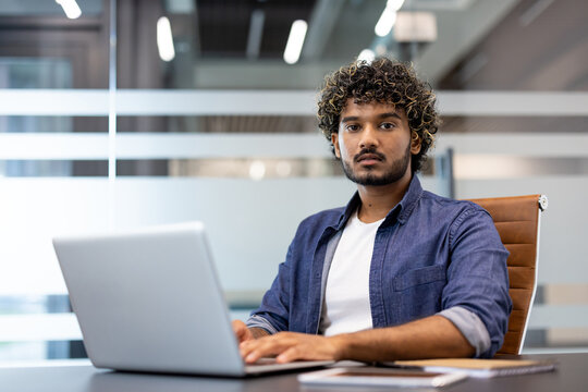Portrait of a young Indian man working in an office, sitting at a desk with a laptop and looking seriously at the camera