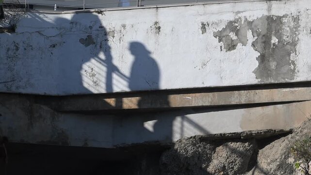 Shadow of a person walking on a bridge near a wall with markings in the afternoon sun on a warm day in an urban setting