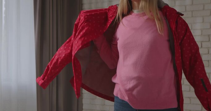 A woman raises her arms as she puts on a pink patterned windbreaker in a bright room with soft lighting. Slow motion.