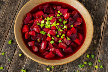 Vegan borscht - a soup made of beetroots, beans, cabbage, carrots, and celery stalks, topped with chopped scallions. Rustic wooden table.