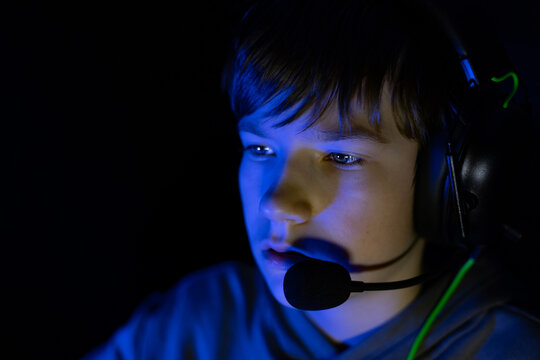 Young boy with headset focused on gaming, illuminated by blue light, showcasing intense concentration and engagement in a digital environment, representing modern gaming culture - Powered by Adobe