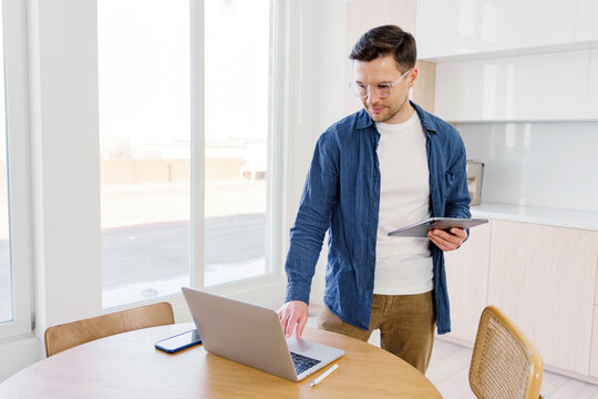 Man works on laptop and holds tablet in home office - Powered by Adobe