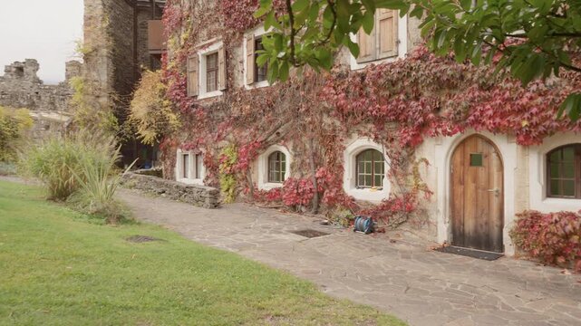 Red virginia creeper leaves cover stone facade of medieval building with arched wooden door. Exterior of historic house with shuttered windows and paved path in autumn garden.