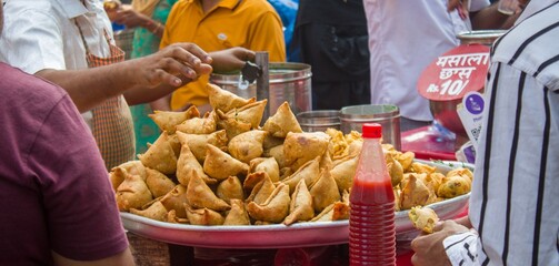 Fototapeta premium Freshly fried Indian samosas stacked on a street food stall with chutney bottle and customers in a busy local market.