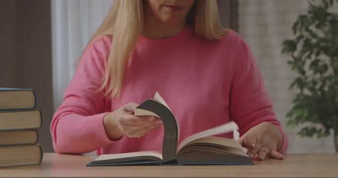 A woman in a pink sweater is sitting at a table, flipping through pages of a book with a stack of books nearby. Slow motion.