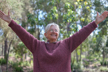 Happy senior woman smiling with arms raised in outdoors. Elderly lady enjoying nature expressing joy and freedom in a forest park on a sunny day. Concept of positivity, active aging, wellbeing